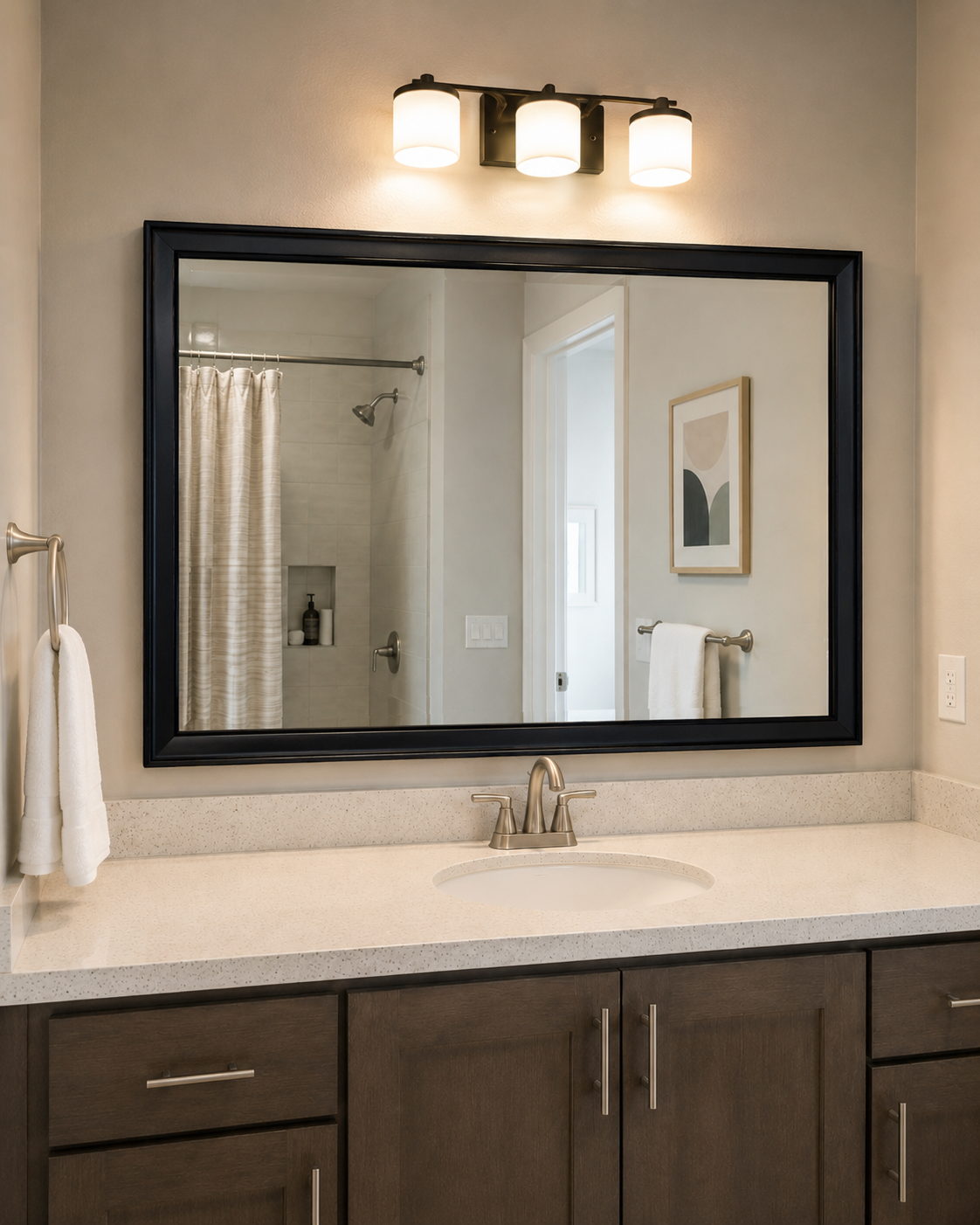 A large rectangular wall mirror with a thick black frame hangs above a white countertop and dark wood bathroom vanity. A three-light wall sconce is mounted above the mirror, and the reflection shows a shower area and framed wall art.