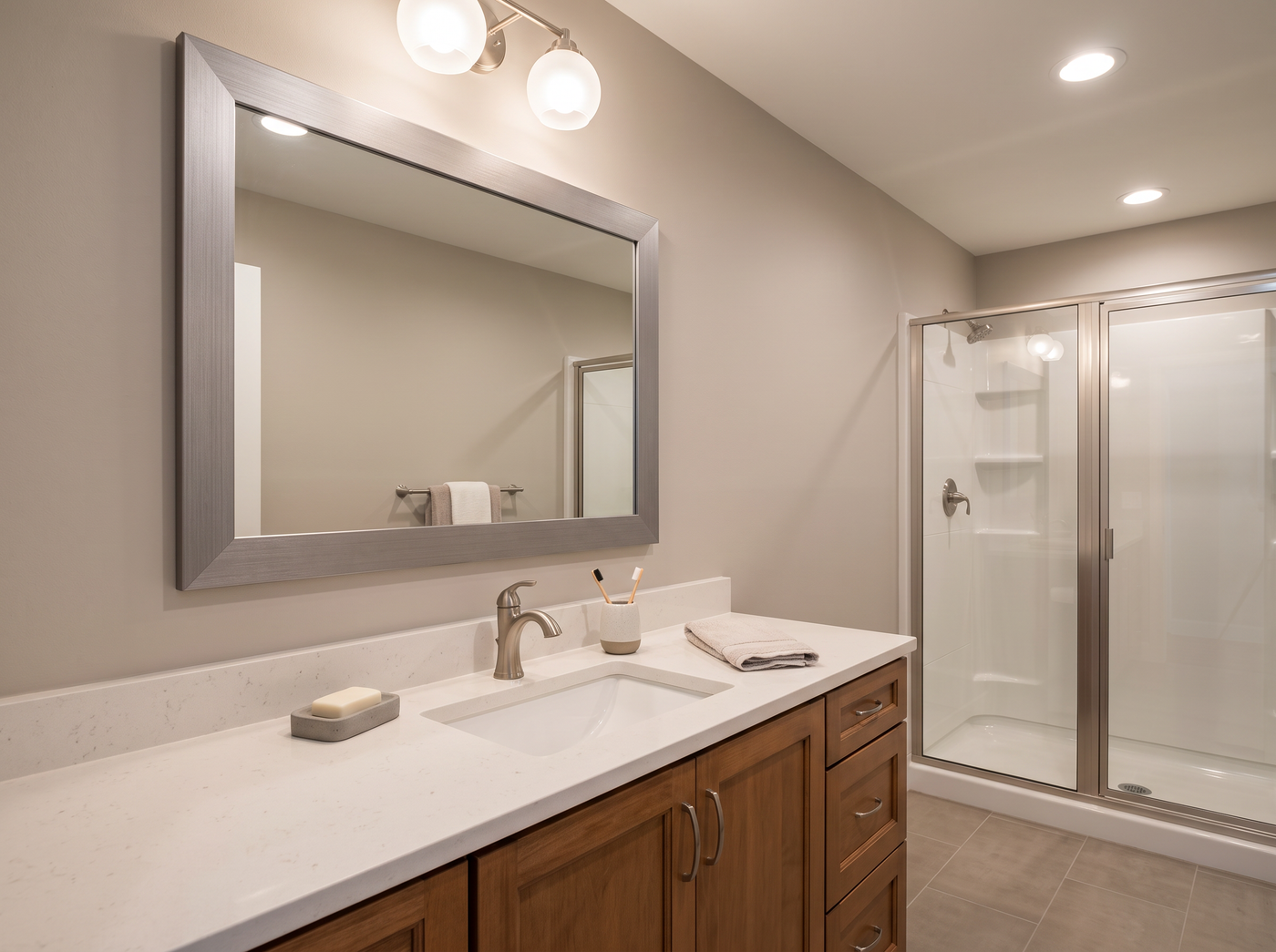 A modern bathroom vanity with a white countertop, white sink, wood cabinet base, large rectangular mirror with a silver frame, and a glass shower door in the background.
